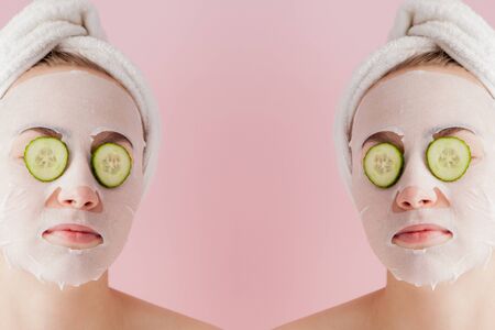 Beautiful young woman is applying a cosmetic tissue mask on a face with cucumber on a pink background.の写真素材