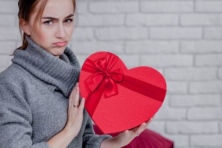 Portrait of a girl in a grey sweater with a heart-shaped red gift box sitting in the apartment, Valentine's day concept, copy space.の写真素材