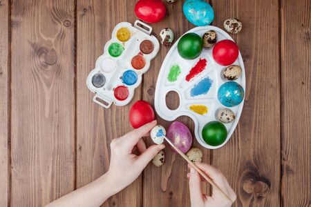 Hand painted Easter eggs, paints and brushes on wooden background. Preparation for the holiday. Girls hands draw a patternの写真素材