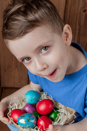 Happy baby boy with a basket of easter eggs on wooden background.