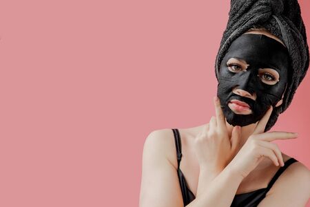 Young woman apply black cosmetic fabric facial mask on pink background. Face peeling mask with charcoal, spa beauty treatment, skincare, cosmetology. Close up.の写真素材