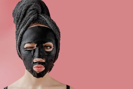 Young woman apply black cosmetic fabric facial mask on pink background. Face peeling mask with charcoal, spa beauty treatment, skincare, cosmetology. Close up.の写真素材