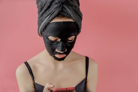 Young woman apply black cosmetic fabric facial mask and phone in hands on pink background. Face peeling mask with charcoal, spa beauty treatment, skincare, cosmetology. Close up.の写真素材