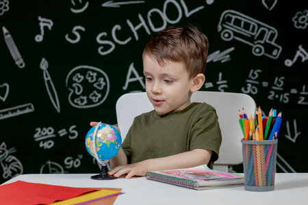 Cute clever boy is sitting at a desk with globe in hand on background with blackboard . Ready for...