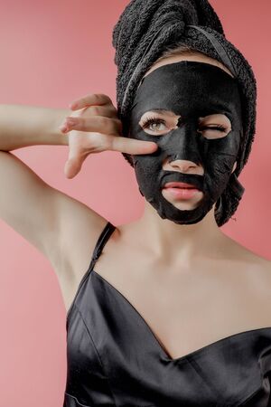 Young woman apply black cosmetic fabric facial mask on pink background. Face peeling mask with charcoal, spa beauty treatment, skincare, cosmetology. Close up.の写真素材