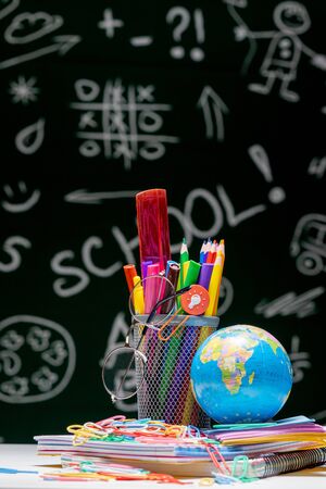 School background with stationery accessories. Books, globe, pencils and various office supplies lying on the desk on a green blackboard background.の写真素材