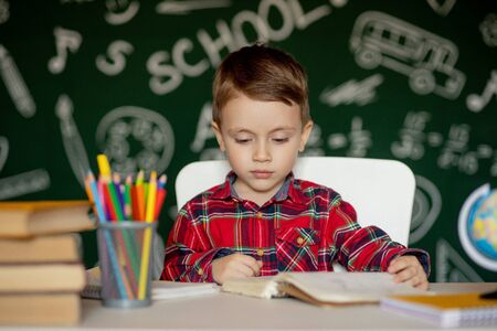Cute clever boy is sitting at a desk with magnifying glass in hand. Child is reading a book with a blackboard on a background. Ready for school. Back to school.の写真素材