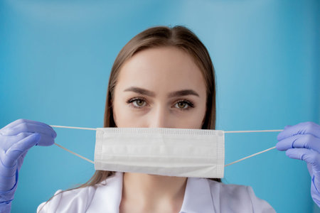 Doctor nurse smiling behind surgeon mask. Closeup portrait of young caucasian woman model on blue...