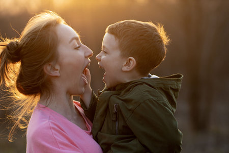 Close up of a lovely mother and her son having fun outdoor. Little cute kid holded by her mom in the arms which is laughing against sunset. Mother's day concept.の写真素材
