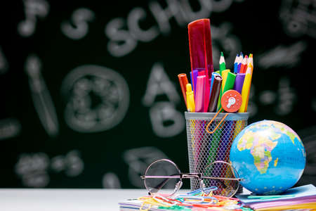 School background with stationery accessories. Books, globe, pencils and various office supplies lying on the desk on a green blackboard backgroundの写真素材