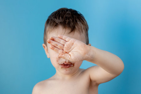 Little boy eating donut chocolate on blue background. Cute happy boy smeared with chocolate around...