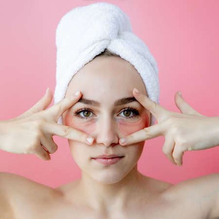Studio shot of satisfied caucasian freckled woman wearing white towel on head, with collagen patches under eyes, standing naked against pink background. Skin care, cosmetic product concept.の写真素材