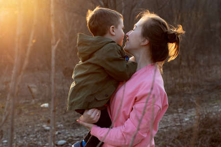 Close up of a lovely mother and her son having fun outdoor. Little cute kid holded by her mom in the arms which is laughing against sunset. Mother's day concept.の写真素材