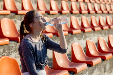 Sporty young attractive girl in sportswear relaxing after hard workout sit and drink water from special sport bottle after running on a stadium.の写真素材