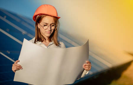 Handsome Architect Woman Examining a Draft Map or Blueprint Project Plan, Worker Activity Looking Out in Photovoltaic Cell Farm or Solar Panels Field.の写真素材