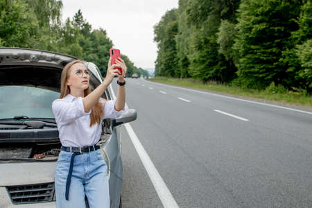 Young woman standing near broken down car with popped up hood having trouble with her vehicle. Waiting for help tow truck or technical support. A woman calls the service center.の写真素材