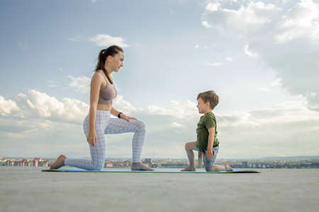 Mother and son doing exercise on the balcony in the background of a city during sunrise or sunset, concept of a healthy lifestyle.の写真素材