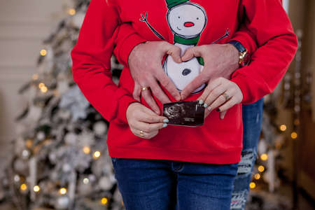 Parents hands keeping ultrasound scan touching and pregnant belly near christmas tree decorated with toys. Cropped, close upの写真素材