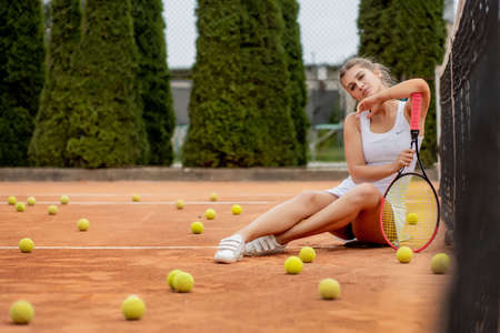 Portrait of beautiful young woman sitting near net in tennis court with ball outdoor. Confident sportswoman resting on tennis courtの写真素材