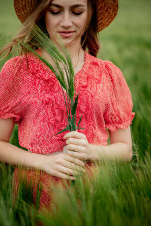 portrait of young woman in dress and hat in green field of barley in countryside. Stylish girl in rustic enjoying peaceful moment in grass in summer. Tranquil rural momentの写真素材