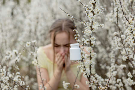 Young girl blowing nose and sneezing in tissue in front of blooming tree. Seasonal allergens affecting people. Beautiful lady has rhinitis.の写真素材