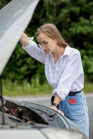 Young woman opening bonnet of broken down car having trouble with her vehicle. Worried woman talking on the phone near broken car. Girl model standing near the broken car calling for auto service.の写真素材