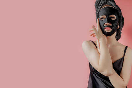 Young woman apply black cosmetic fabric facial mask on pink background. Face peeling mask with charcoal, spa beauty treatment, skincare, cosmetology. Close up.の写真素材
