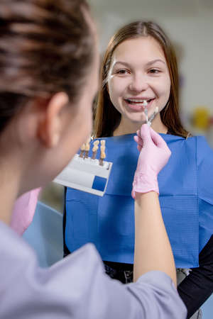 Close up portrait of beautiful young lady sitting in dental chair while stomatologist hands in sterile gloves holding tooth samplesの写真素材