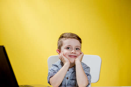 Happy child boy sitting at table with laptop and preparing to school on the yellow background. Online learning. Online schoolの写真素材