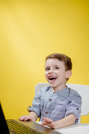 Happy child boy sitting at table with laptop and preparing to school on the yellow background....
