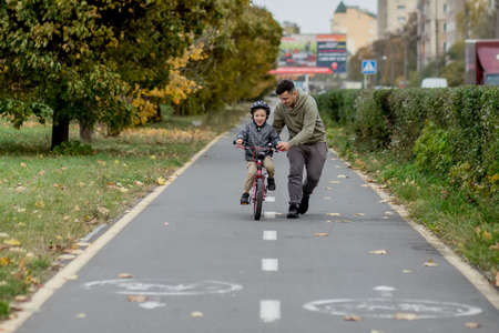 Father teaches his son to ride a bicycle on the bike path in the park. The father is holding a bicycle and the son is sitting on it.の写真素材