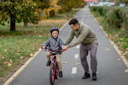 Father teaches his son to ride a bicycle on the bike path in the park. The father is holding a bicycle and the son is sitting on it.の写真素材