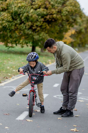 Father teaches his son to ride a bicycle on the bike path in the park. The father is holding a bicycle and the son is sitting on it.の写真素材