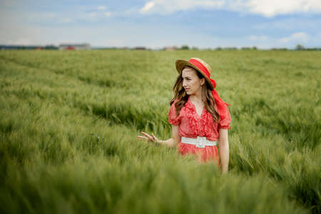 Young woman in dress and hat in green field of barley in countryside. Tranquil rural momentの写真素材