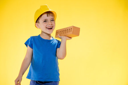 Boy in a construction helmet holds a brick in his hands on yellow background.