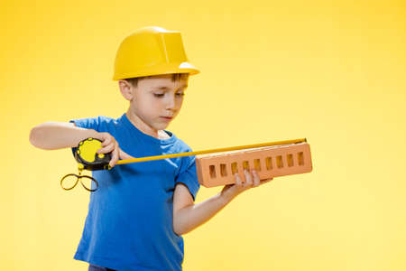 Boy in a construction helmet holds a brick in his hands and measures it with a tape measure staiding in studio on yellow backgroundの写真素材