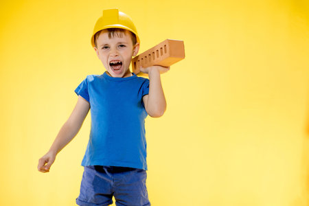 Boy in a construction helmet holds a brick in his hands on yellow background.の写真素材