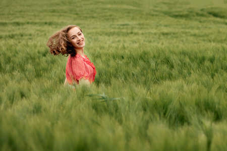 Young woman in red dress and hat walking in green field of barley in countryside. Tranquil rural moment.の写真素材