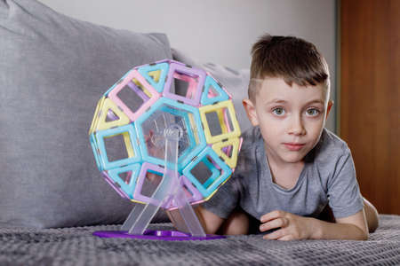 A boy in a gray T-shirt sits on a bed near a white wall and plays with a Ferris wheel, which he designed with a magnetic constructor. Creativity development. magnetic constructor.の写真素材