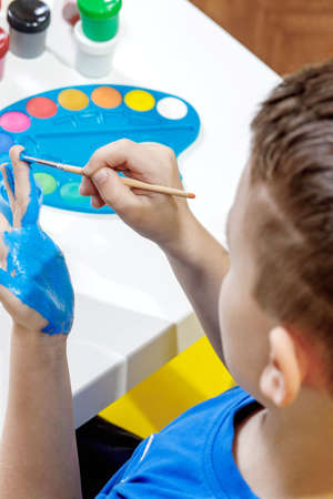 A little preschooler sits at a table, holding a brush in his hand and painting paints his palm. preschooler developmentの写真素材