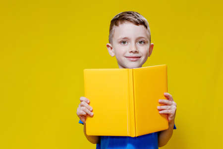 Positive preschool boy in an blueT-shirt holding an open yellow copybook on yellow background.の写真素材