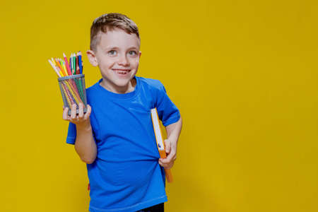 Smiling happy schooler in blue t-shirt holding multicolored pencils and book on yellow background.の写真素材