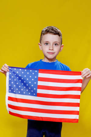 Little smiling boy holding an American flag on a yellow background. Patriotism, independence day, flag day concept. Remembrance day.の写真素材