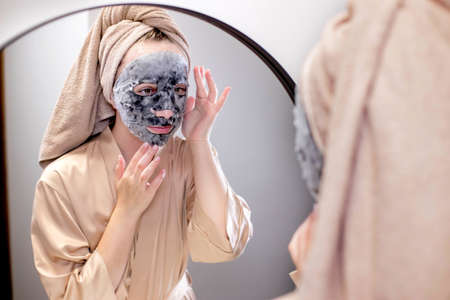Young woman applying face mask in bathroom and smiling. Beautiful woman in front of a mirror doing beauty treatmentsの写真素材