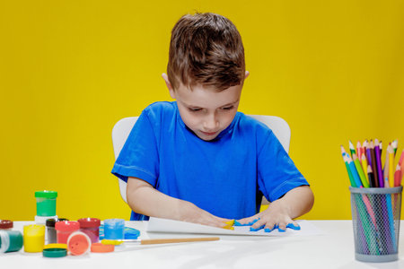 A little preschooler is sitting at a table, drawing with his palms on a sheet of paper. Colorful...