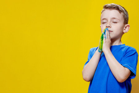Ukrainian boy prays for Ukraine. Children against war. A boy in a blue T-shirt on a yellow background folded his hands painted in the Ukrainian flag for prayer.の写真素材