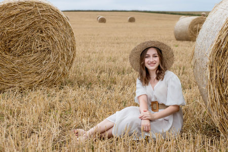 A young woman in a dress and hat posing in a field near hay bales. A woman near a haystack in a...