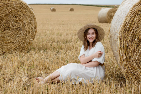 A young woman in a dress and hat posing in a field near hay bales. A woman near a haystack in a field. Summer photoset.の写真素材