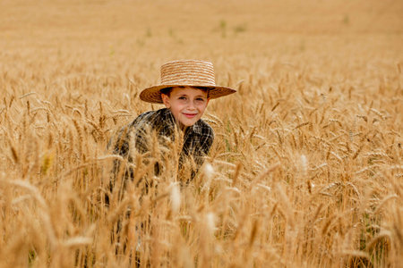 A smiling little farmer boy in a plaid shirt and straw hat poses for a photo in a wheat field. Heir of farmers.の写真素材