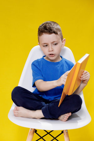 Little cheerful blond green-eyed boy 5-6 years old in a stylish blue T-shirt holding book and reading on yellow wall background, children's studio portrait.の写真素材